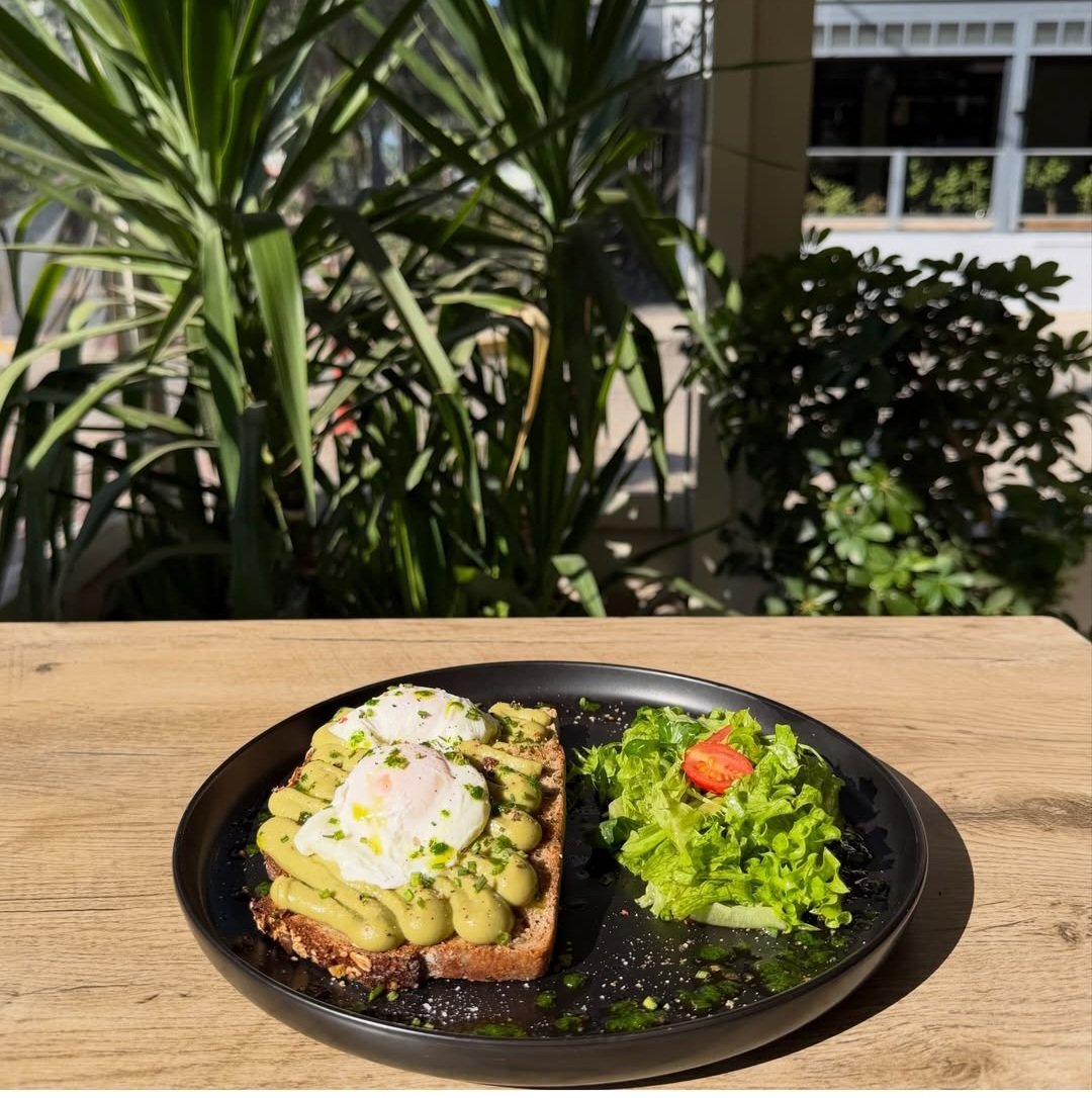 Avocado toast with poached egg and fresh green salad on black plate at outdoor cafe with plants and city building visible in background