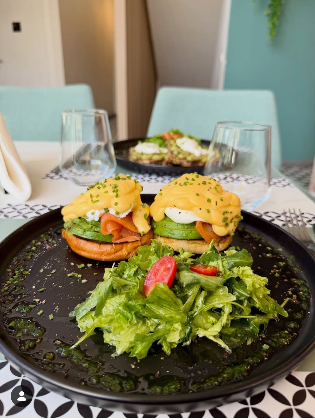 Two turmeric-dusted sandwiches with lettuce and egg on a black plate, served with greens and pickled ginger in a modern restaurant setting