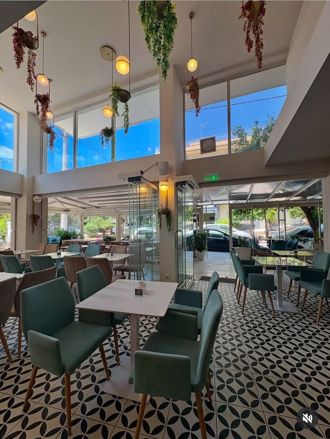 Modern restaurant interior with green upholstered chairs, white tables, hanging plants, pendant lights, and floor-to-ceiling windows overlooking outdoor patio