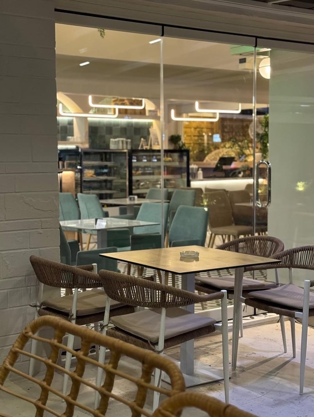 Modern outdoor patio seating area with woven chairs, wooden tables, and floor cushions facing a contemporary glass storefront